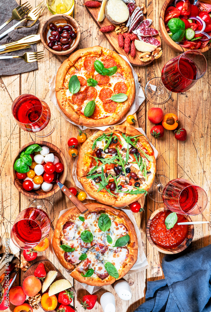 Pizza party table. Top view glasses with red wine, rustic wooden table with hot pizzas, italian appetizers, salads, cheese, fruits and berries. Family lunch with fast food. Vertical image Pizza party table. Top view glasses with red wine, rustic wooden table with hot pizzas, italian appetizers, salads, cheese, fruits and berries. Family lunch with fast food. Vertical image