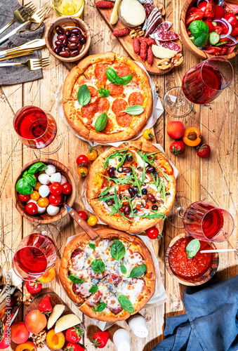 Pizza party table. Top view glasses with red wine, rustic wooden table with hot pizzas, italian appetizers, salads, cheese, fruits and berries. Family lunch with fast food. Vertical image
