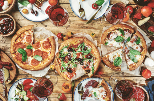 Pizza party table. Top view glasses with red wine, rustic wooden table with hot pizzas, italian appetizers, salads, cheese, fruits and berries. Family lunch with fast food