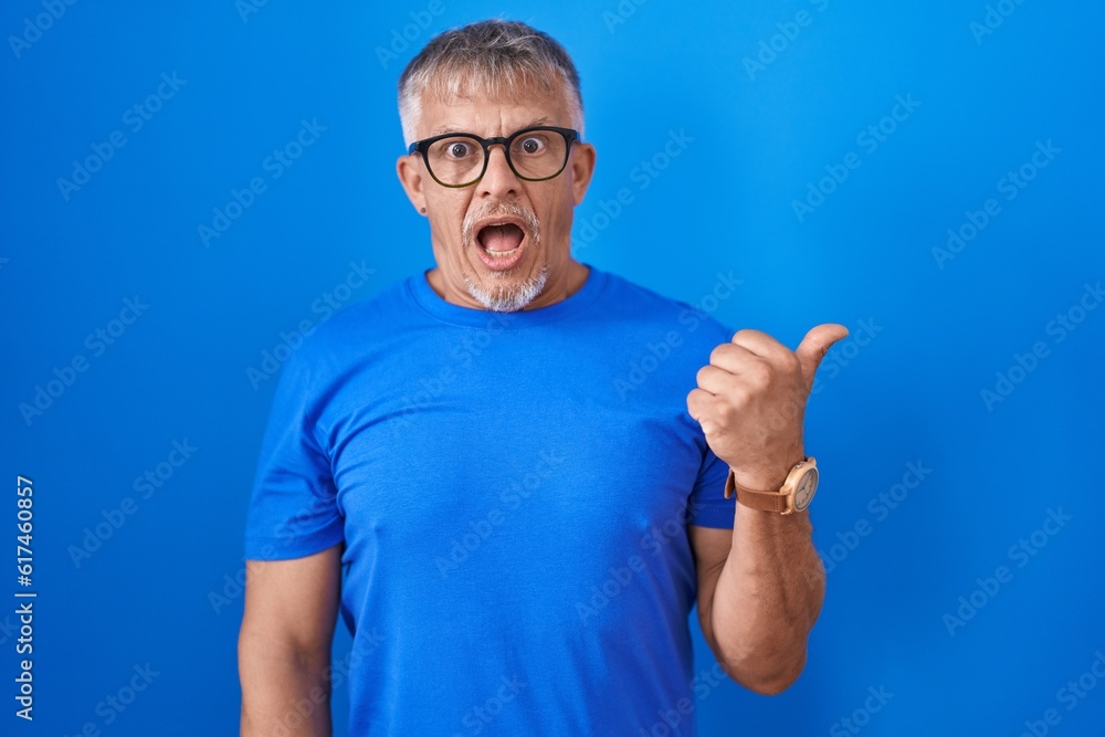 Hispanic man with grey hair standing over blue background surprised pointing with hand finger to the side, open mouth amazed expression. Hispanic man with grey hair standing over blue background surprised pointing with hand finger to the side, open mouth amazed expression.