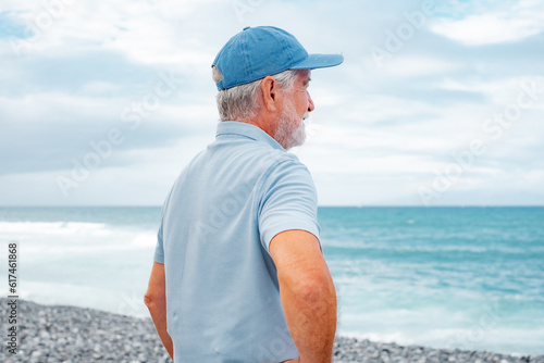 Rear view of senior man in casual polo shirt standing at the beach looking at horizon over sea, pensioner male with cap enjoying free time and retirement