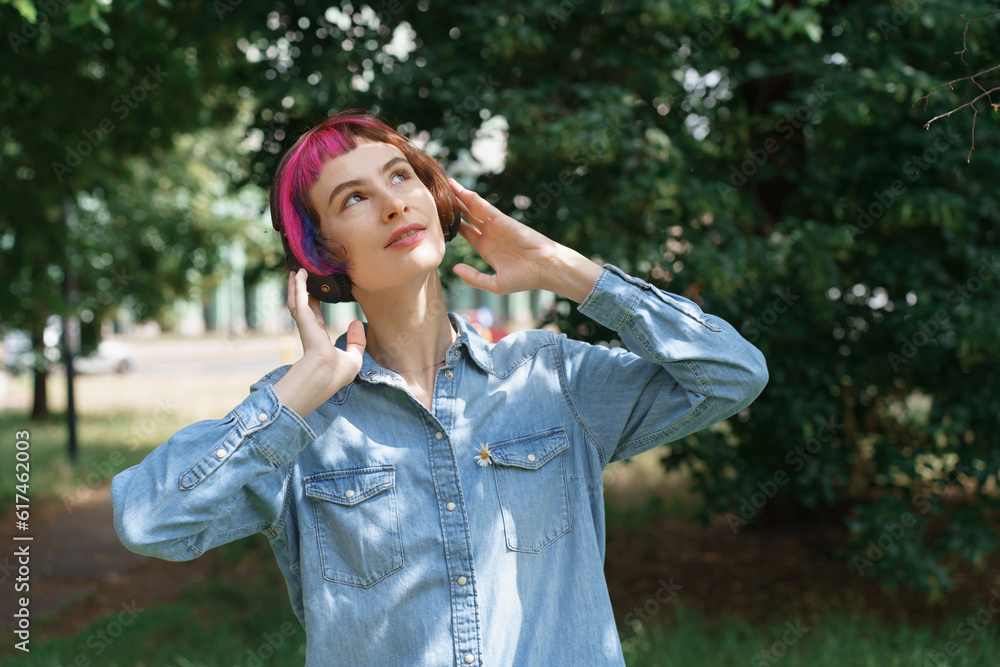 A girl with pink hair with headphones listens to music in the park in summer