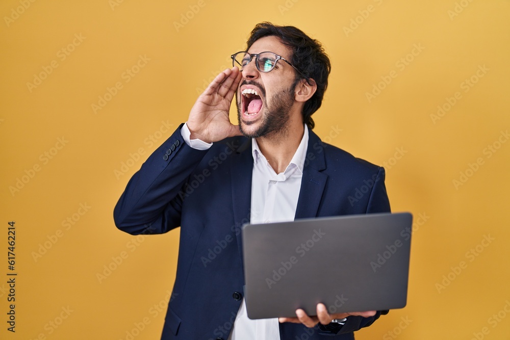 Handsome latin man working using computer laptop shouting and screaming loud to side with hand on mouth. communication concept. Handsome latin man working using computer laptop shouting and screaming loud to side with hand on mouth. communication concept.