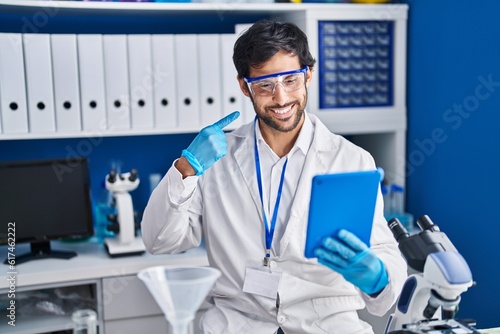Handsome latin man working at scientist laboratory using tablet smiling happy pointing with hand and finger