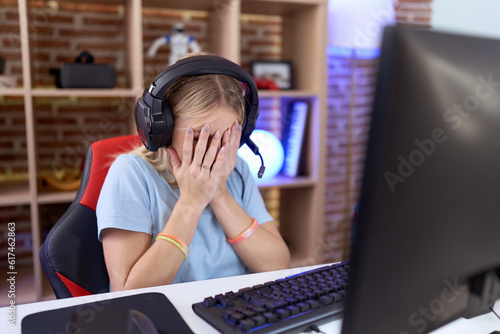 Young caucasian woman playing video games wearing headphones with sad expression covering face with hands while crying. depression concept.