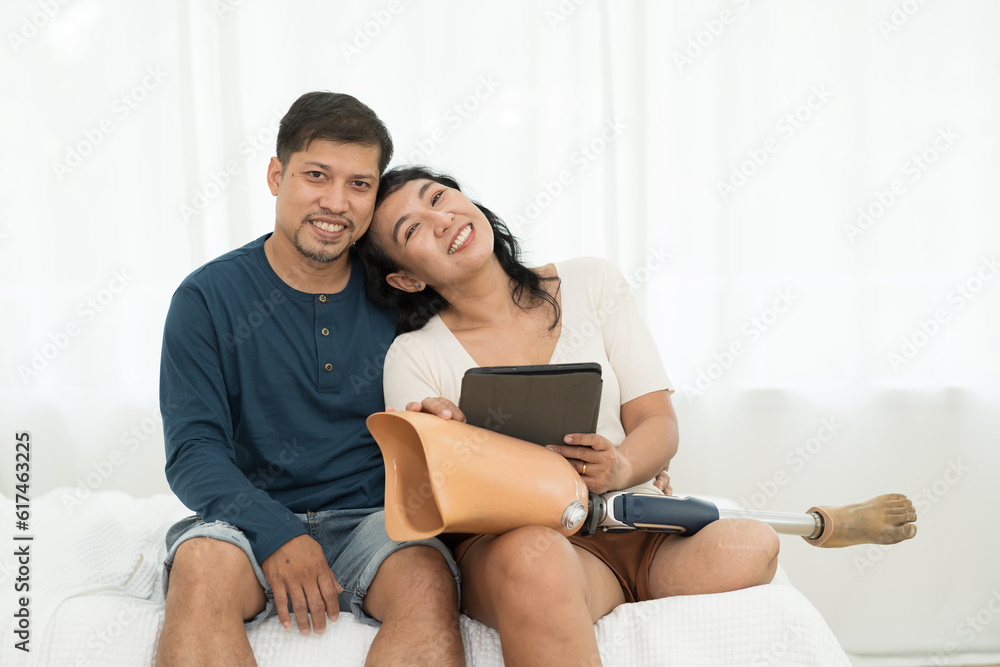 Happy smiling Asian couple embrace together and holding prosthetic leg sitting in bedroom at home. Asian woman with prosthetic leg hugging with her husband at home Happy smiling Asian couple embrace together and holding prosthetic leg sitting in bedroom at home. Asian woman with prosthetic leg hugging with her husband at home