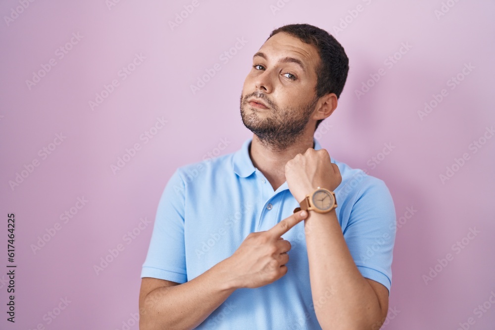 Hispanic man standing over pink background in hurry pointing to watch time, impatience, looking at the camera with relaxed expression Hispanic man standing over pink background in hurry pointing to watch time, impatience, looking at the camera with relaxed expression