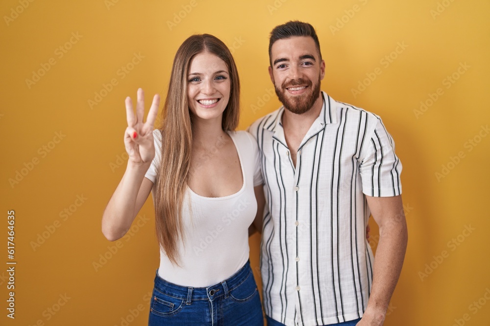Φωτογραφία Young couple standing over yellow background showing and pointing up with fingers number three while smiling confident and happy