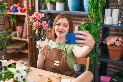 Young redhead woman florist make selfie by smartphone holding plant at flower shop