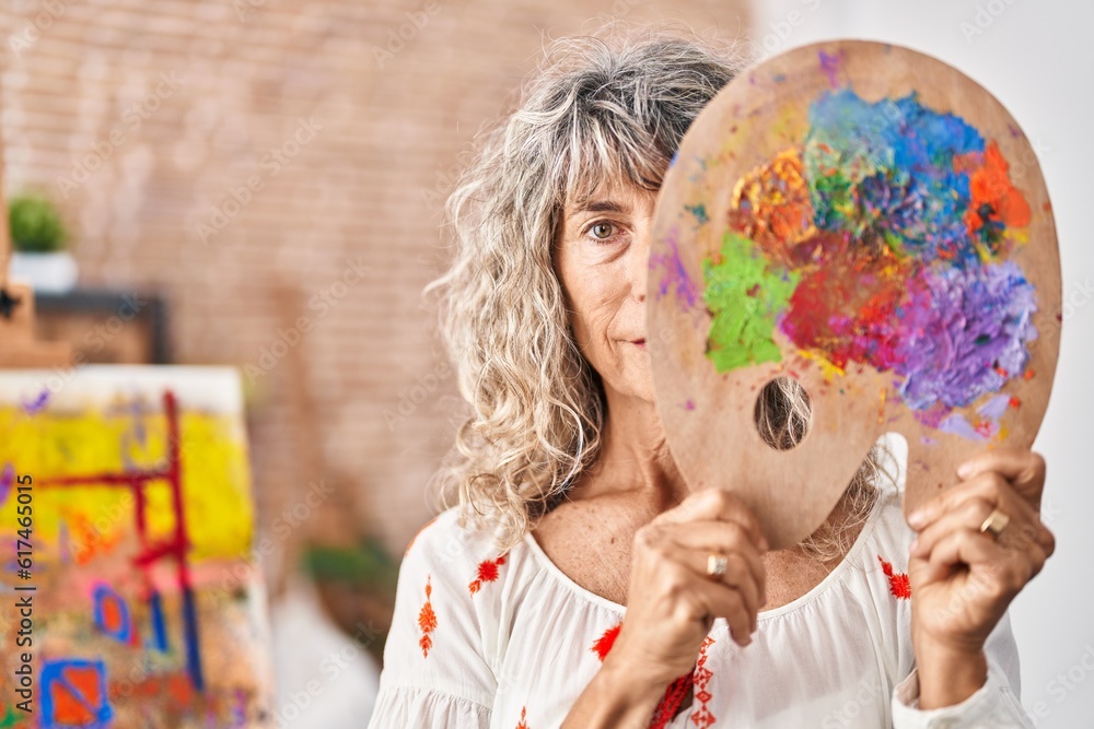 Fototapeta premium Middle age woman holding painter palette covering face relaxed with serious expression on face. simple and natural looking at the camera.