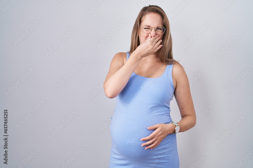 Young pregnant woman standing over white background smelling something stinky and disgusting, intolerable smell, holding breath with fingers on nose. bad smell Young pregnant woman standing over white background smelling something stinky and disgusting, intolerable smell, holding breath with fingers on nose. bad smell