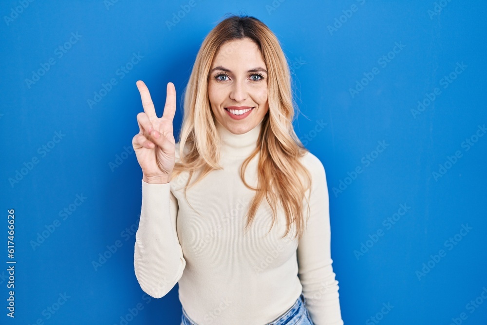 Young caucasian woman standing over blue background showing and pointing up with fingers number two while smiling confident and happy. Young caucasian woman standing over blue background showing and pointing up with fingers number two while smiling confident and happy.