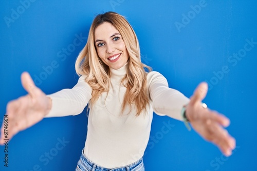 Young caucasian woman standing over blue background looking at the camera smiling with open arms for hug. cheerful expression embracing happiness. © Krakenimages.com