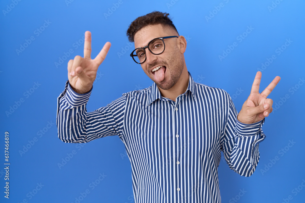 Hispanic man with beard wearing glasses smiling with tongue out showing fingers of both hands doing victory sign. number two. Hispanic man with beard wearing glasses smiling with tongue out showing fingers of both hands doing victory sign. number two.