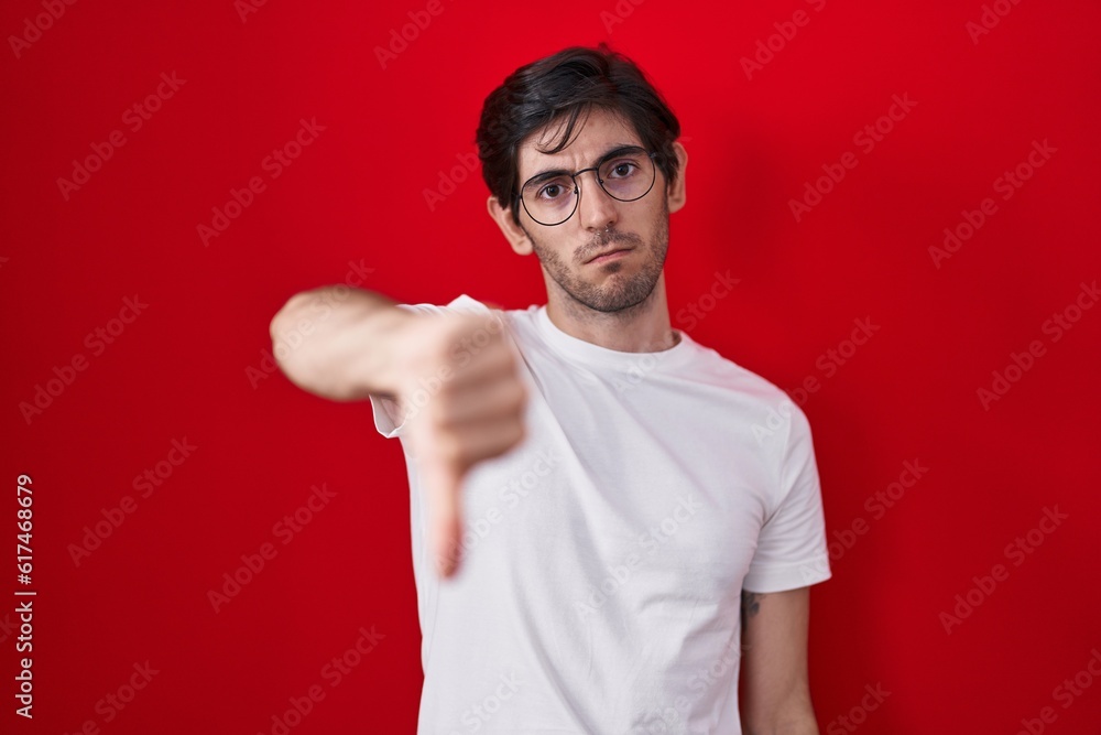 Young hispanic man standing over red background looking unhappy and angry showing rejection and negative with thumbs down gesture. bad expression. Young hispanic man standing over red background looking unhappy and angry showing rejection and negative with thumbs down gesture. bad expression.