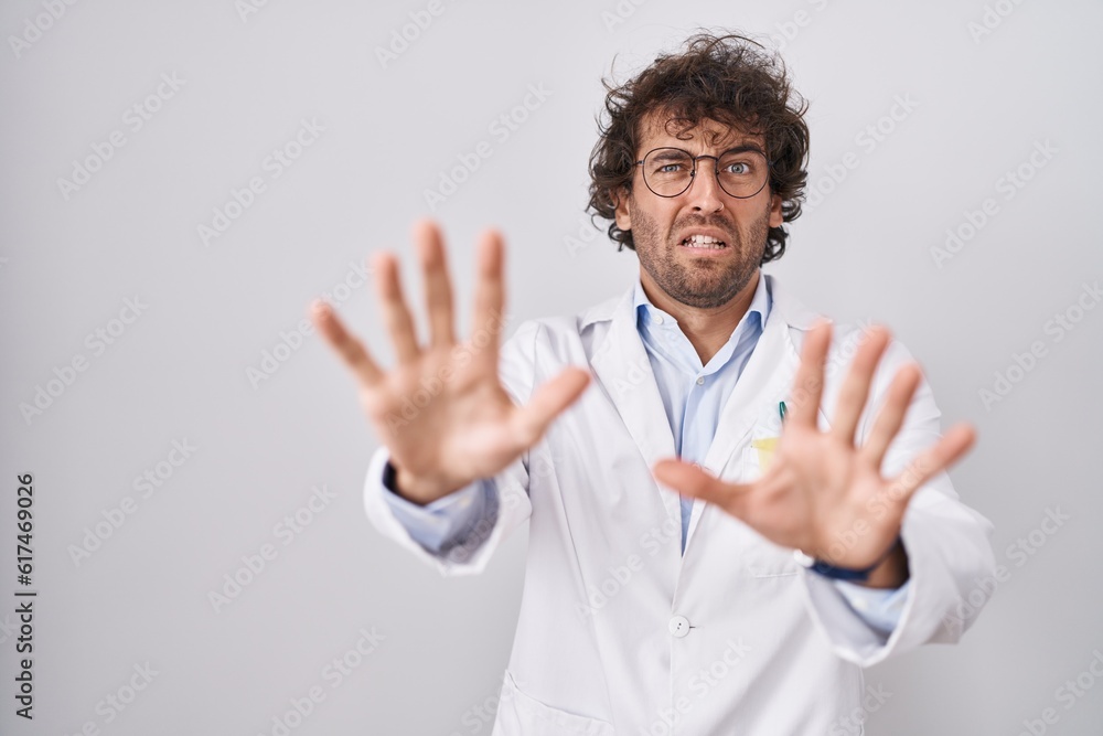 Hispanic young man wearing doctor uniform afraid and terrified with fear expression stop gesture with hands, shouting in shock. panic concept. Hispanic young man wearing doctor uniform afraid and terrified with fear expression stop gesture with hands, shouting in shock. panic concept.