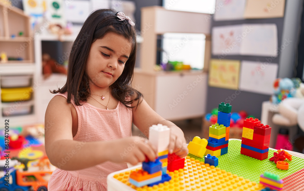 Fototapeta premium Adorable hispanic girl playing with construction blocks sitting on table at kindergarten
