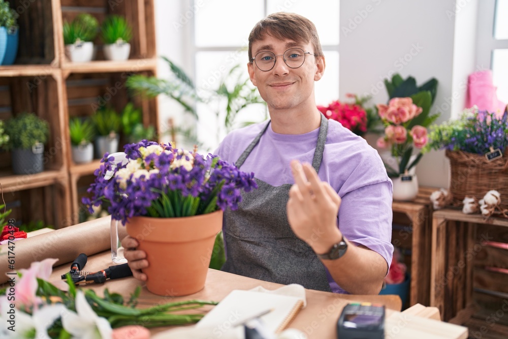 Caucasian blond man working at florist shop beckoning come here gesture with hand inviting welcoming happy and smiling