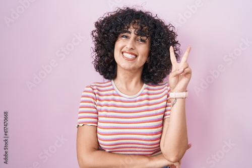 Young middle east woman standing over pink background smiling with happy face winking at the camera doing victory sign with fingers. number two. © Krakenimages.com