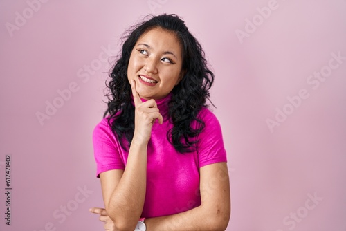 Young asian woman standing over pink background with hand on chin thinking about question, pensive expression. smiling and thoughtful face. doubt concept.