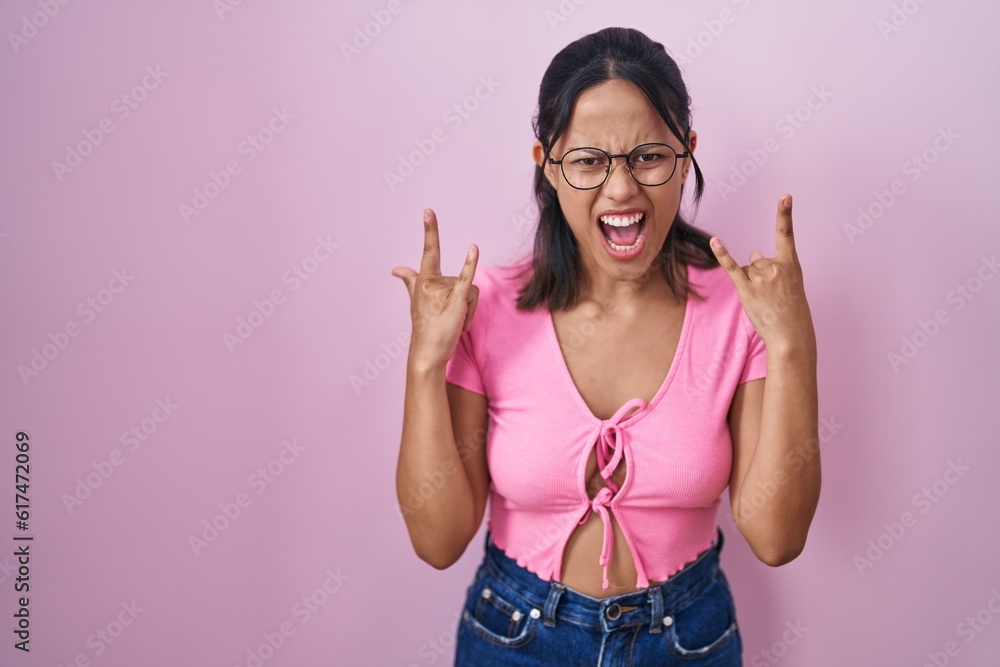 Hispanic young woman standing over pink background wearing glasses shouting with crazy expression doing rock symbol with hands up. music star. heavy concept. Hispanic young woman standing over pink background wearing glasses shouting with crazy expression doing rock symbol with hands up. music star. heavy concept.
