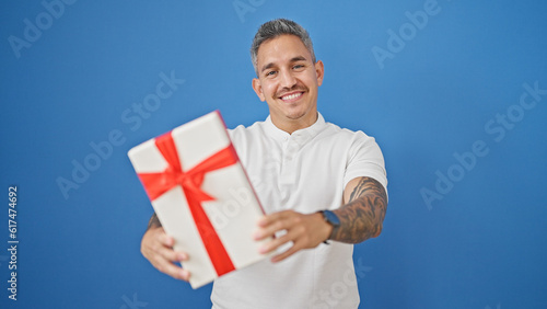 Young hispanic man smiling confident holding gift over isolated blue background