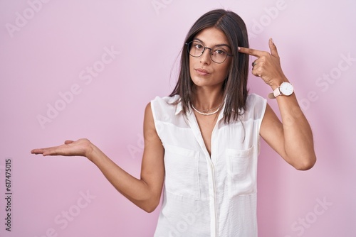 Brunette young woman standing over pink background wearing glasses confused and annoyed with open palm showing copy space and pointing finger to forehead. think about it.