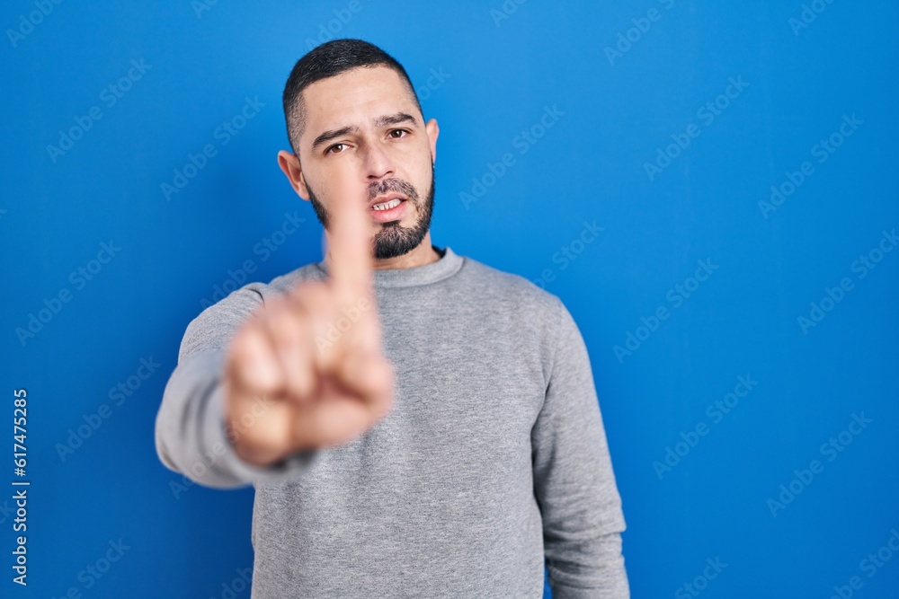 Hispanic man standing over blue background pointing with finger up and angry expression, showing no gesture Hispanic man standing over blue background pointing with finger up and angry expression, showing no gesture