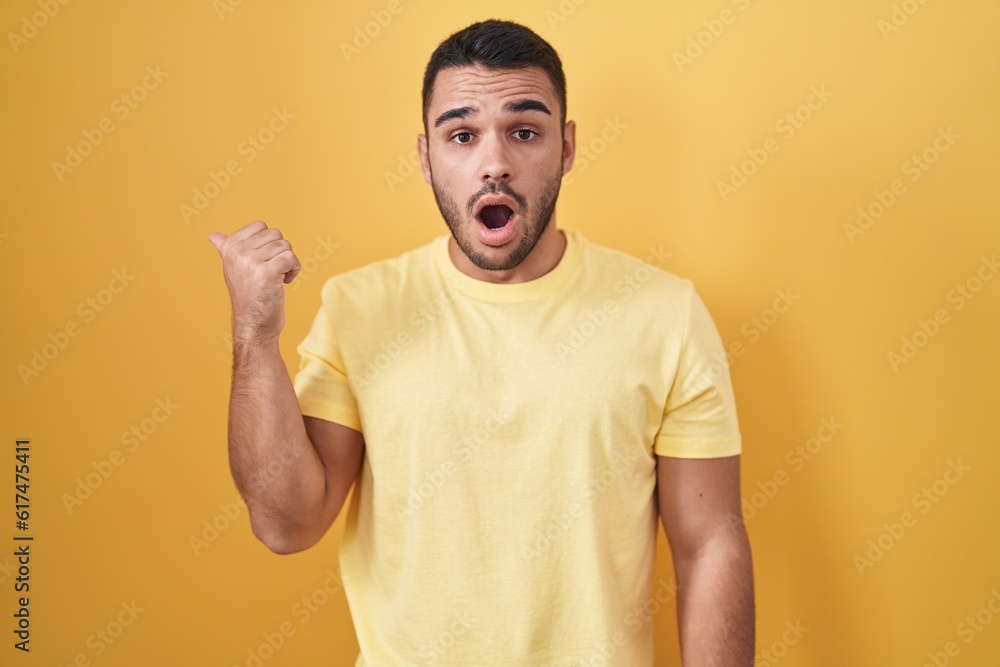 Young hispanic man standing over yellow background surprised pointing with hand finger to the side, open mouth amazed expression. Young hispanic man standing over yellow background surprised pointing with hand finger to the side, open mouth amazed expression.