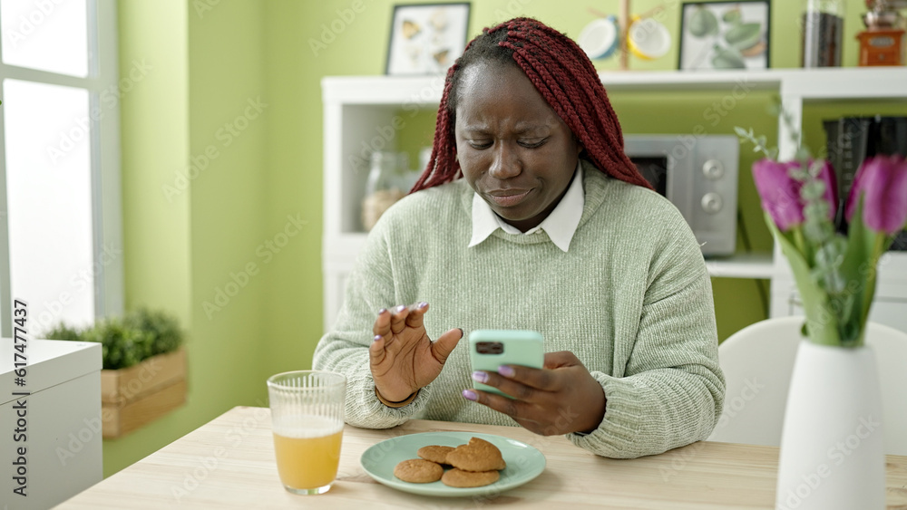 African woman with braided hair eating cookies using smartphone at dinning room African woman with braided hair eating cookies using smartphone at dinning room