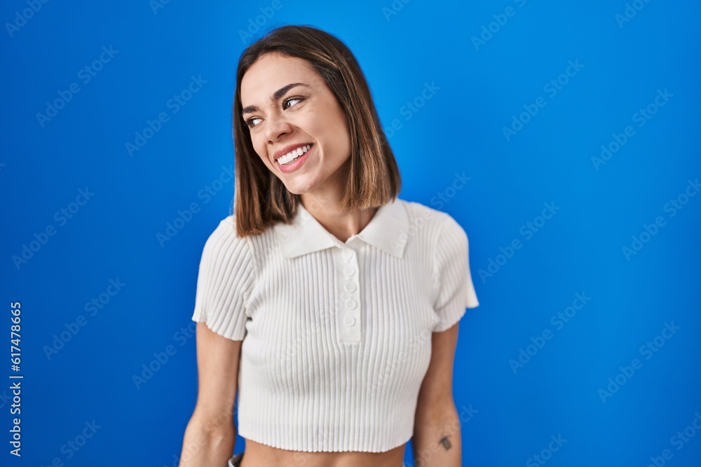 Hispanic woman standing over blue background looking away to side with smile on face, natural expression. laughing confident. Hispanic woman standing over blue background looking away to side with smile on face, natural expression. laughing confident.