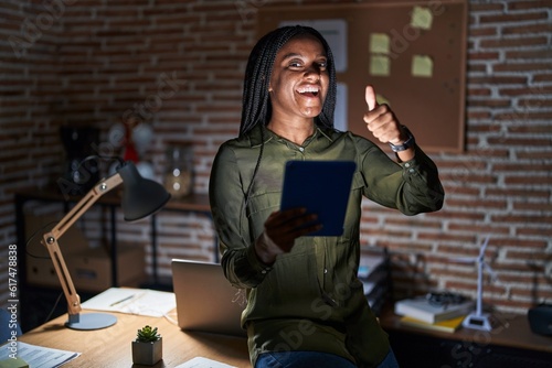 Young african american with braids working at the office at night approving doing positive gesture with hand, thumbs up smiling and happy for success. winner gesture.