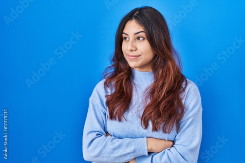 Hispanic young woman standing over blue background looking to the side with arms crossed convinced and confident
