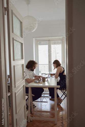 Married couple freelancers sit at table in living room with laptops and working from home office