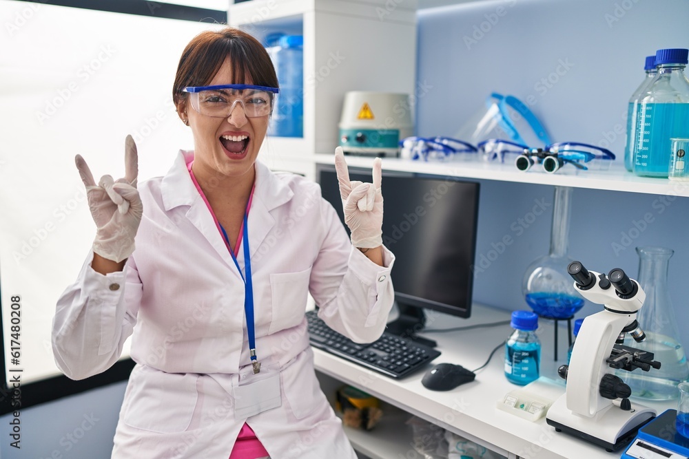 Young brunette woman working at scientist laboratory shouting with crazy expression doing rock symbol with hands up. music star. heavy concept. Young brunette woman working at scientist laboratory shouting with crazy expression doing rock symbol with hands up. music star. heavy concept.