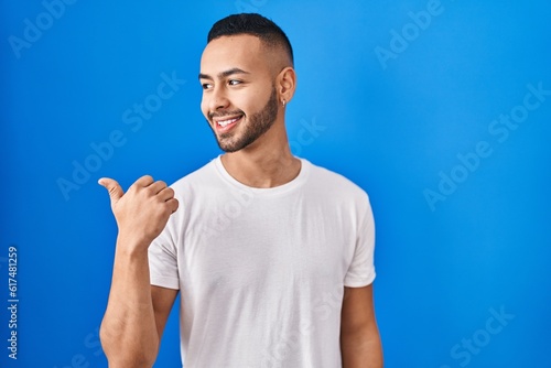Young hispanic man standing over blue background smiling with happy face looking and pointing to the side with thumb up.