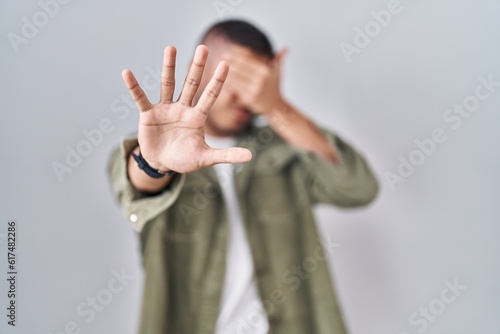 Young hispanic man standing over isolated background covering eyes with hands and doing stop gesture with sad and fear expression. embarrassed and negative concept.