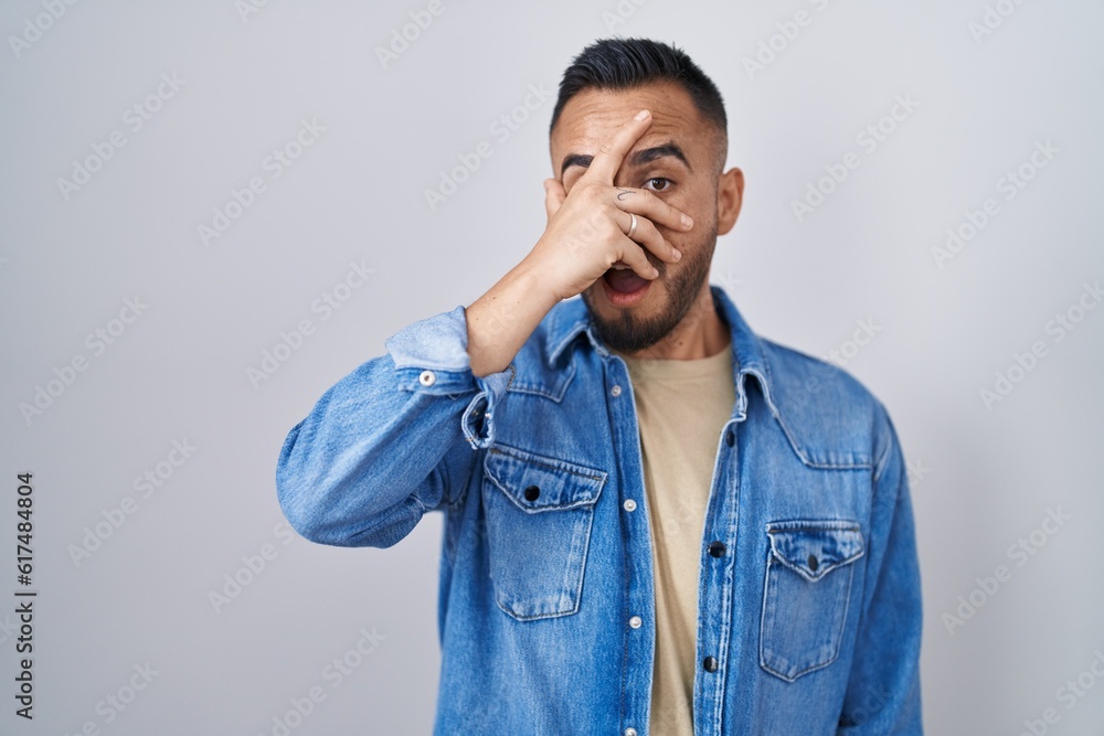 Young hispanic man standing over isolated background peeking in shock covering face and eyes with hand, looking through fingers with embarrassed expression. Young hispanic man standing over isolated background peeking in shock covering face and eyes with hand, looking through fingers with embarrassed expression.