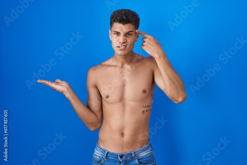 Young hispanic man standing shirtless over blue background confused and annoyed with open palm showing copy space and pointing finger to forehead. think about it.