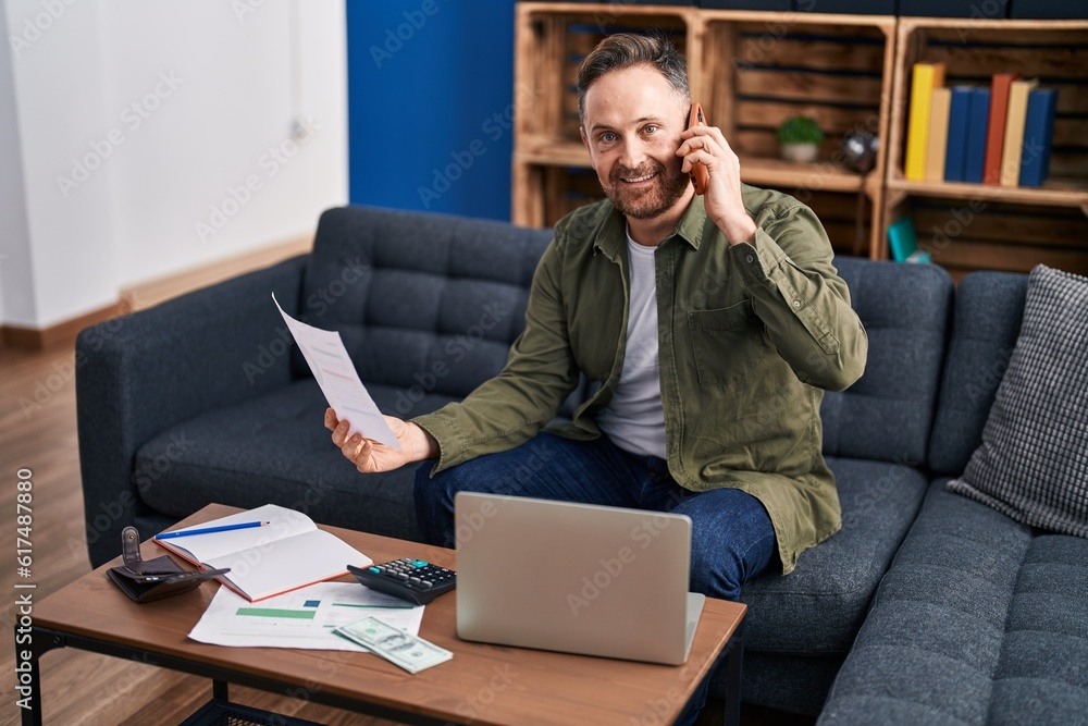 Young caucasian man talking on smartphone reading document at home Young caucasian man talking on smartphone reading document at home