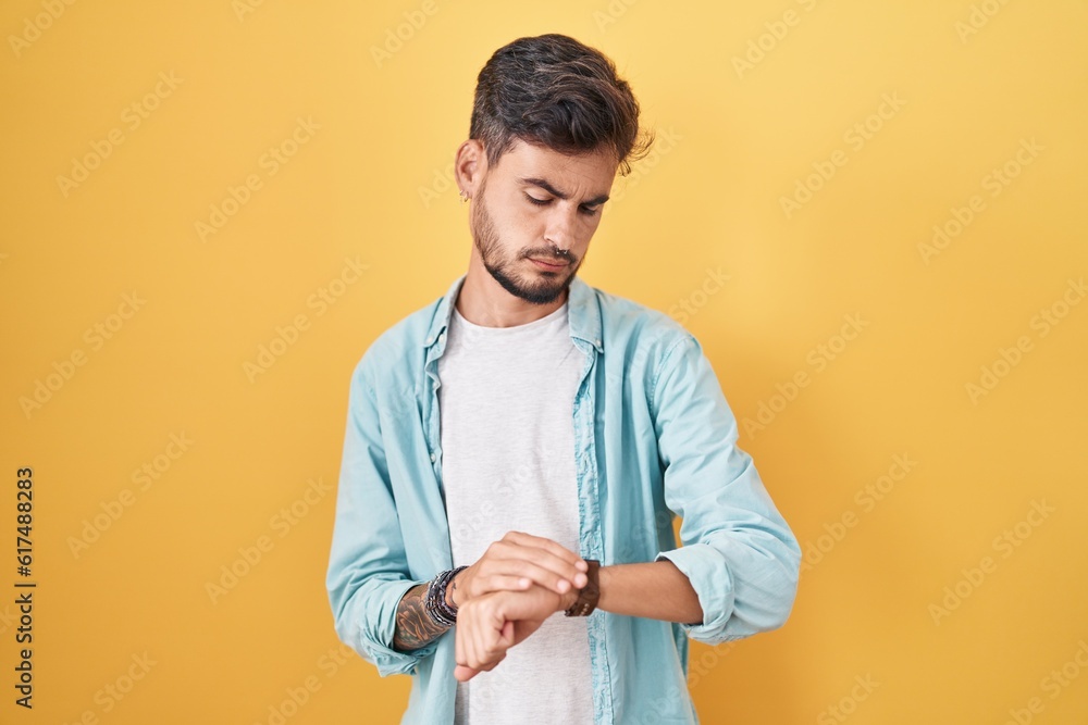 Young hispanic man with tattoos standing over yellow background checking the time on wrist watch, relaxed and confident Young hispanic man with tattoos standing over yellow background checking the time on wrist watch, relaxed and confident