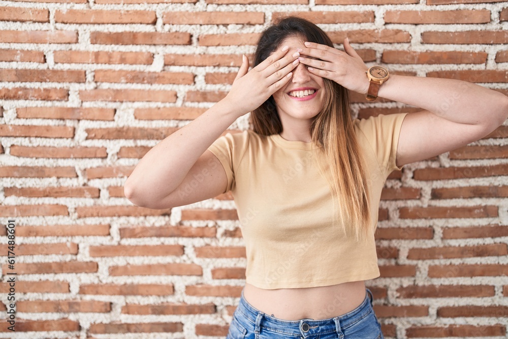 Young brunette woman standing over bricks wall covering eyes with hands smiling cheerful and funny. blind concept. Young brunette woman standing over bricks wall covering eyes with hands smiling cheerful and funny. blind concept.