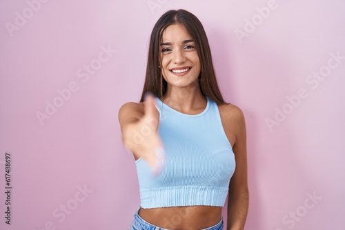 Young brunette woman standing over pink background smiling friendly offering handshake as greeting and welcoming. successful business.