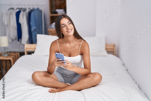 Young beautiful hispanic woman using smartphone sitting on bed at bedroom