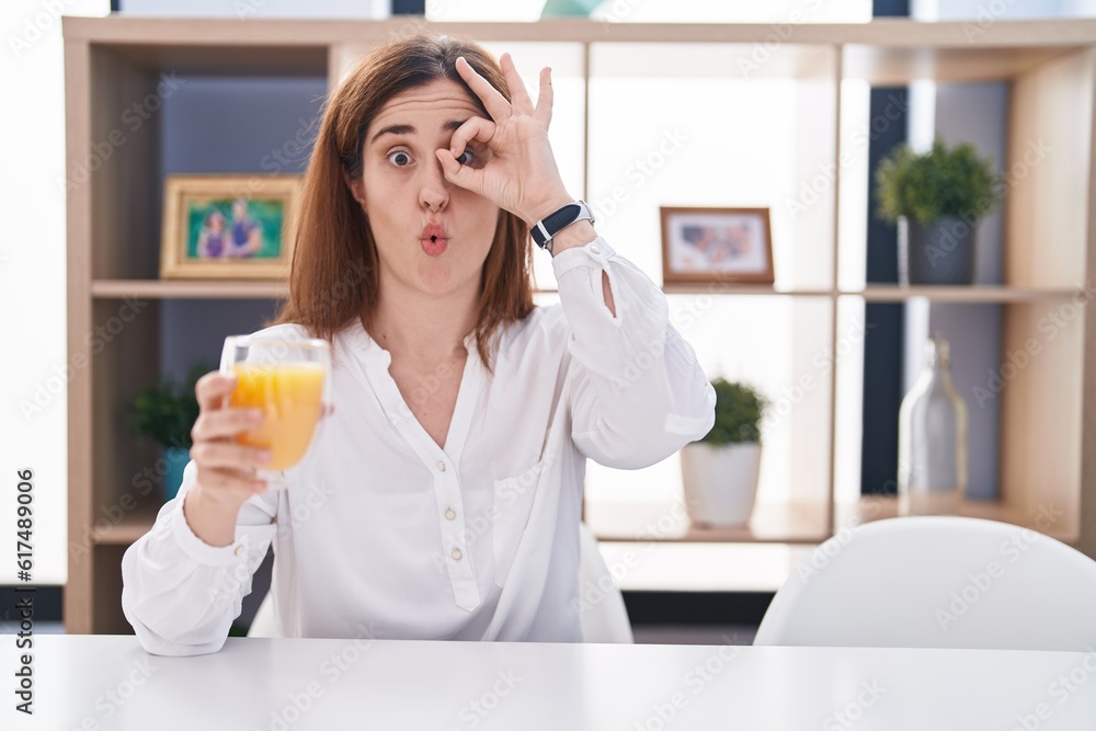 Brunette woman drinking glass of orange juice doing ok gesture shocked with surprised face, eye looking through fingers. unbelieving expression. Brunette woman drinking glass of orange juice doing ok gesture shocked with surprised face, eye looking through fingers. unbelieving expression.