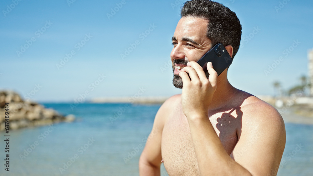 Young hispanic man tourist talking on smartphone standing shirtless at the beach