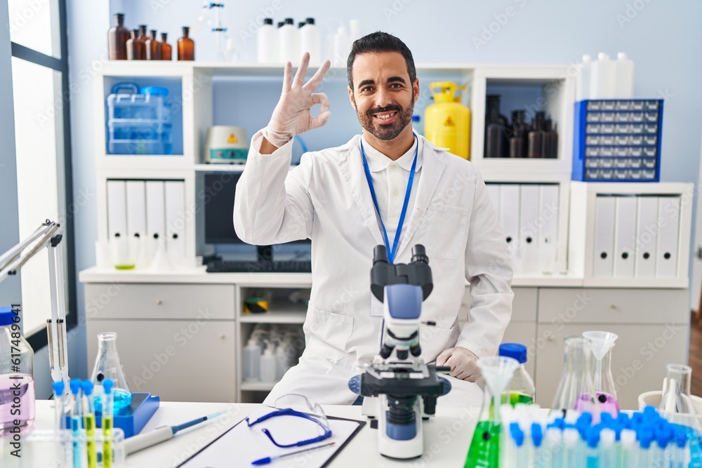 Fototapeta premium Young hispanic man with beard working at scientist laboratory smiling positive doing ok sign with hand and fingers. successful expression.