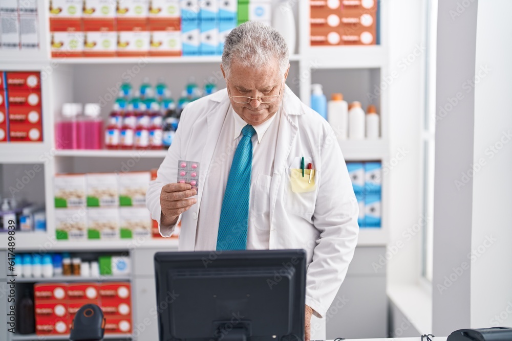 Middle age grey-haired man pharmacist using computer holding pills tablet at pharmacy Middle age grey-haired man pharmacist using computer holding pills tablet at pharmacy