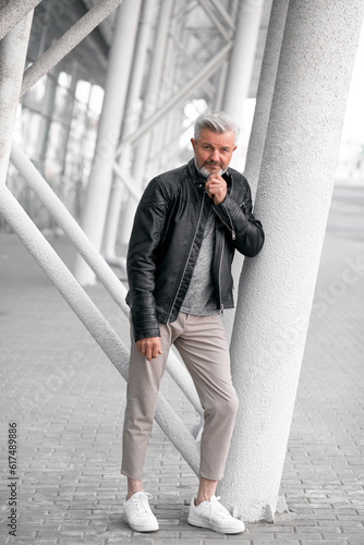 Portrait of a contented older gentleman amidst beautiful white urban background. Mid-Adult stylish white bearded man, in a leather jacket, staying with straight sight.
