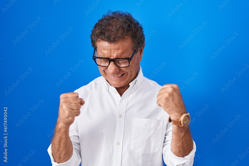 Middle age hispanic man standing over blue background excited for success with arms raised and eyes closed celebrating victory smiling. winner concept. Middle age hispanic man standing over blue background excited for success with arms raised and eyes closed celebrating victory smiling. winner concept.